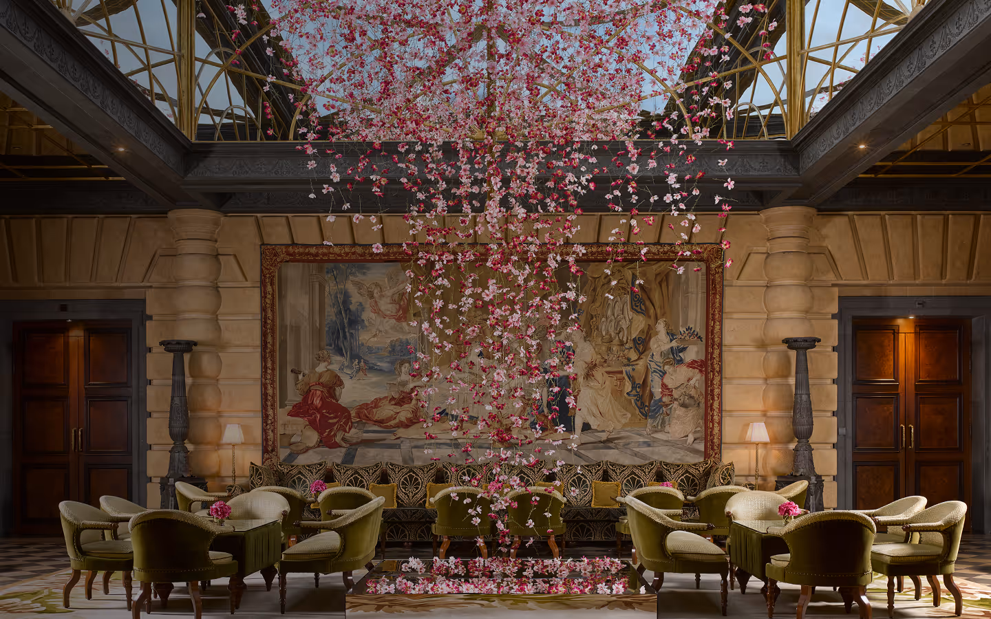 Elegant room with velvet green chairs and tables, a large classical tapestry, and hanging pink and white floral installation from a glass ceiling.