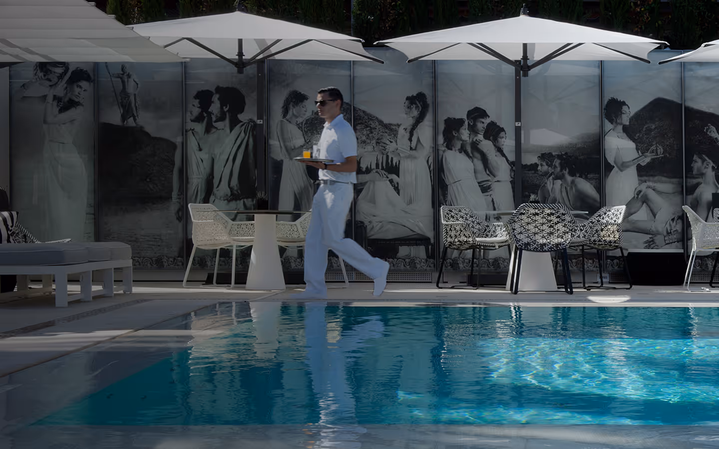 Man in white uniform carrying a tray with drinks, walking beside a swimming pool with outdoor tables, chairs, and umbrellas, and a large black-and-white mural in the background.