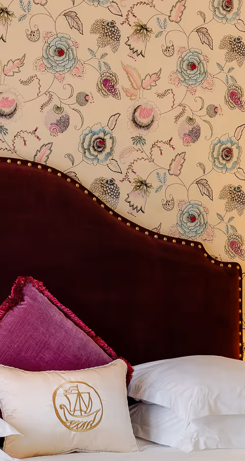 Bed with a dark burgundy velvet headboard decorated with brass studs, against floral patterned wallpaper, featuring white pillows and a decorative pillow with a gold emblem.