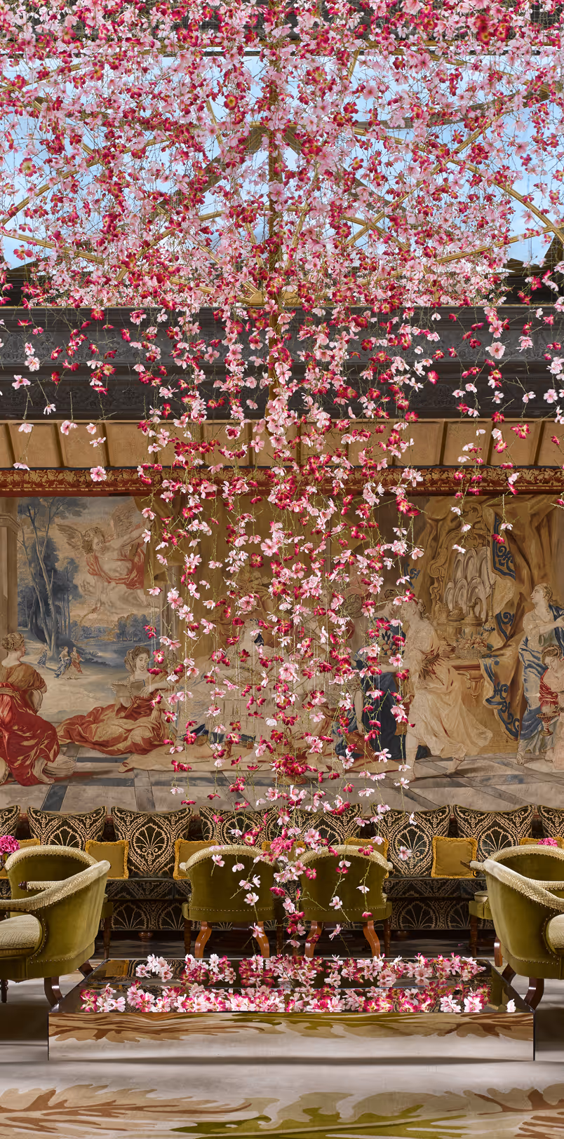 Elegant room with green velvet chairs, a decorative floral ceiling installation of pink flowers, and a detailed classical wall tapestry.