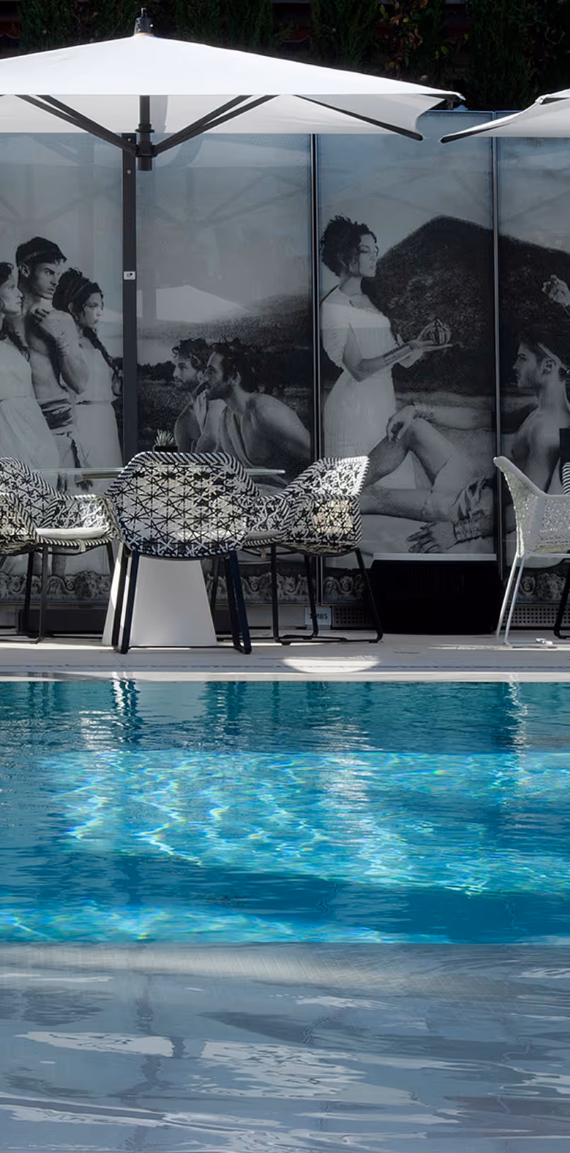 Outdoor poolside area with blue water, patterned chairs around tables, white umbrellas, and a wall mural depicting a classical scene with people.