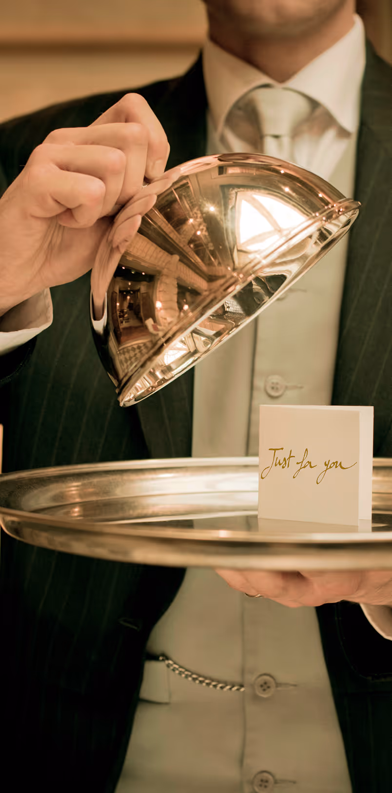 Waiter lifting a shiny cloche off a tray revealing a card that reads 'Just for you'.