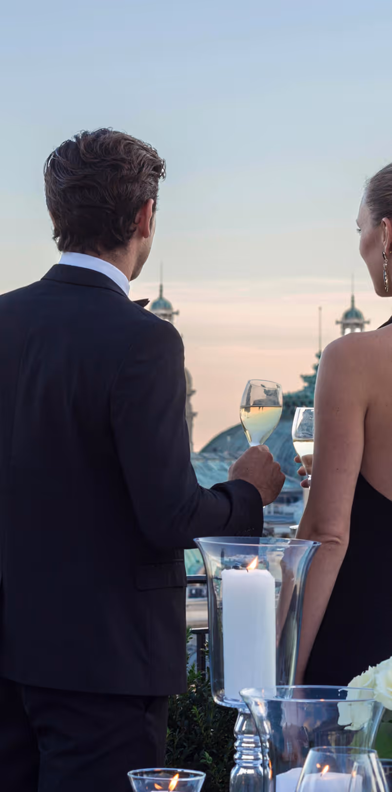 Man and woman dressed elegantly, holding wine glasses and looking at a cityscape during sunset.