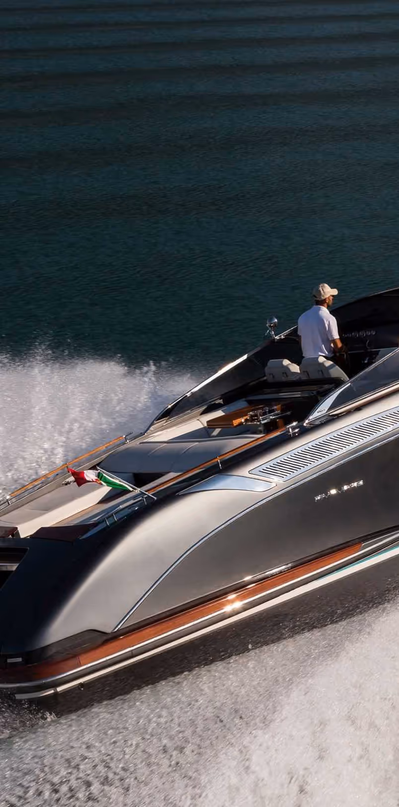 Man driving a sleek, silver motorboat on dark blue water with a wake trailing behind.