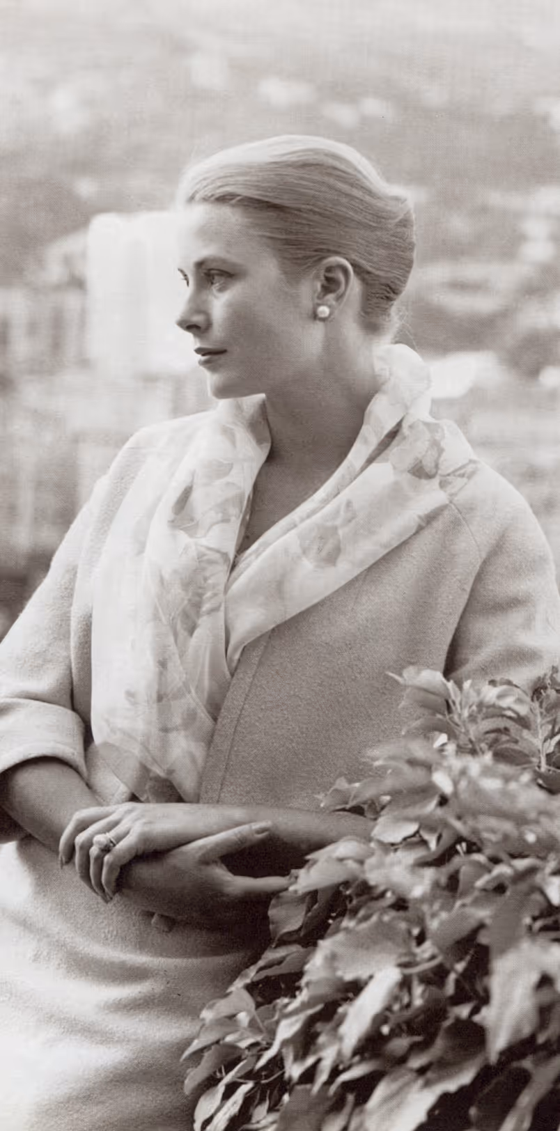 Black and white portrait of a woman with hair in a bun, wearing earrings and a shawl, looking to the left with hands resting on a leafy bush.
