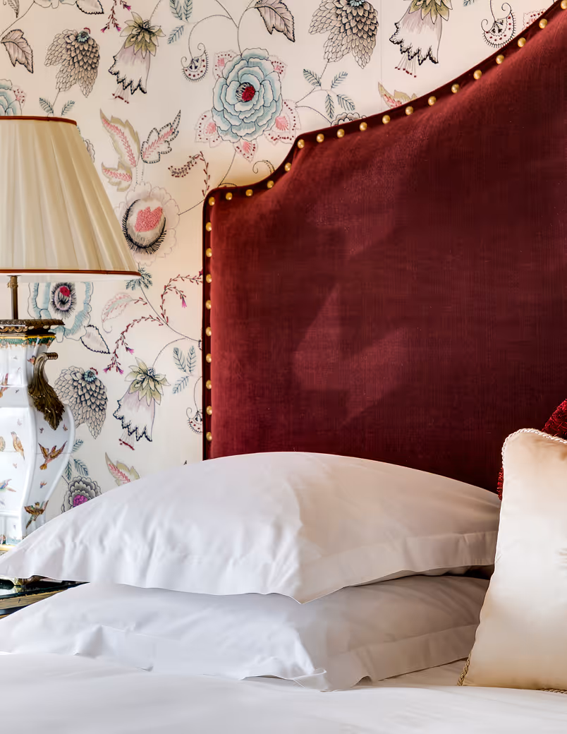 Close-up of a bed with white pillows, a red velvet headboard with gold studs, floral wallpaper, and a decorative lamp.