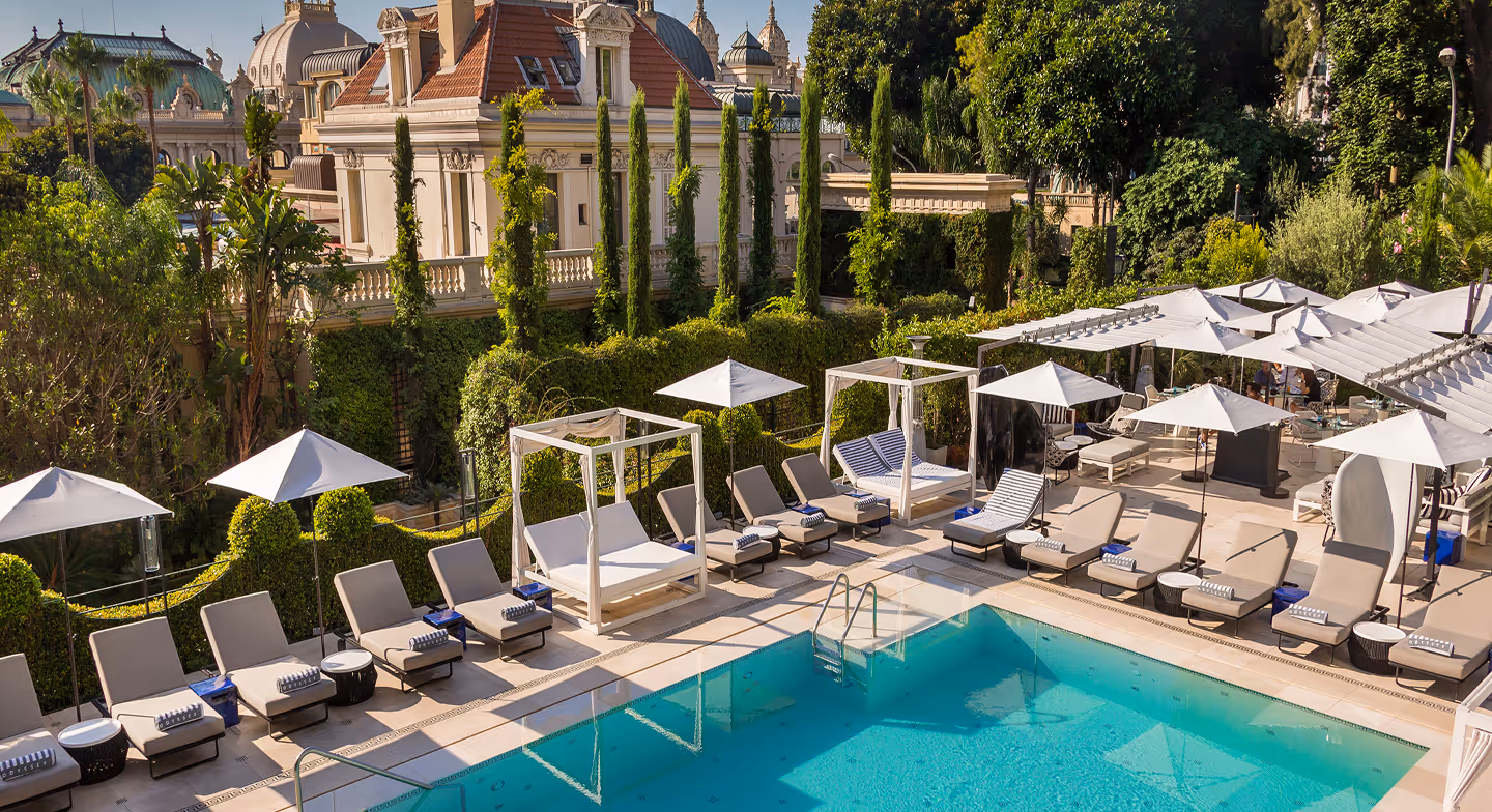 Outdoor pool area with blue water surrounded by beige lounge chairs, white umbrellas, and greenery, with classical architecture in the background.