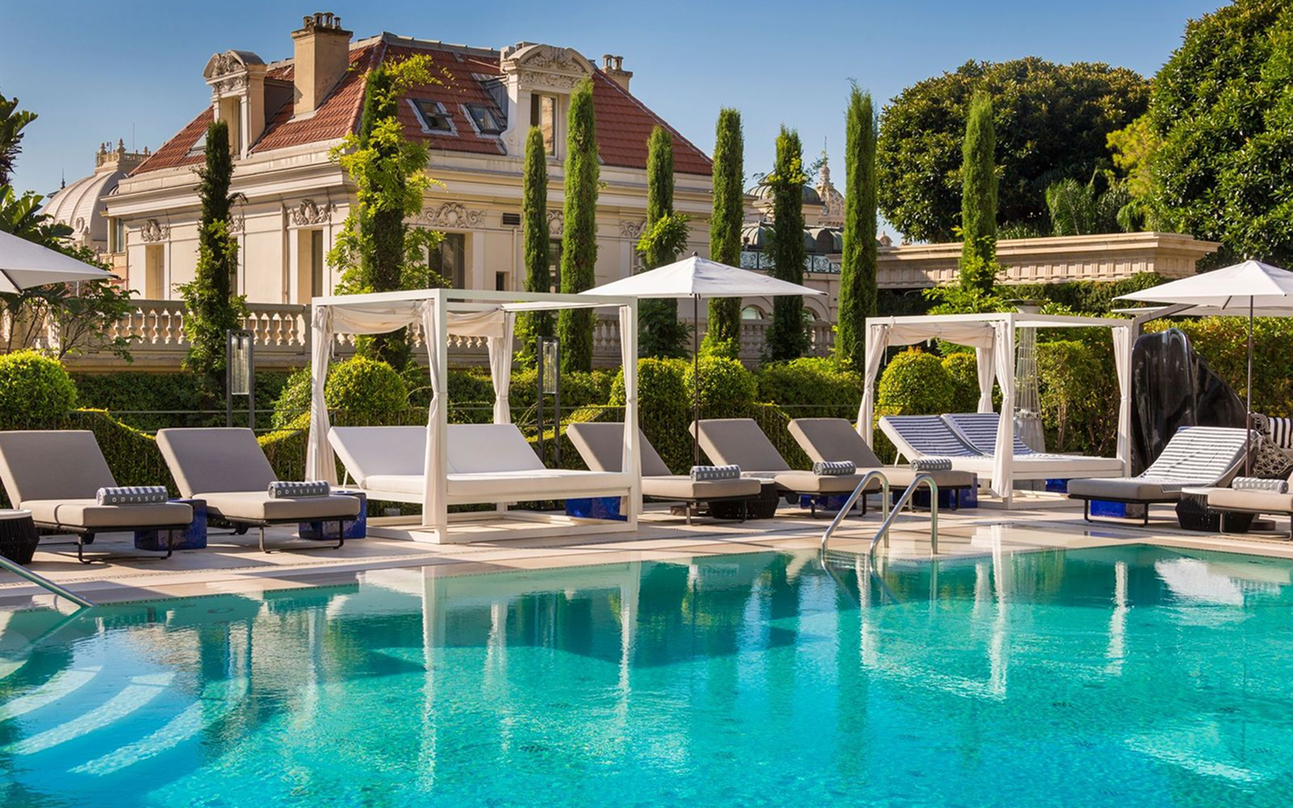 Outdoor swimming pool with turquoise water surrounded by lounge chairs, cabanas, umbrellas, and greenery in front of a classic building.