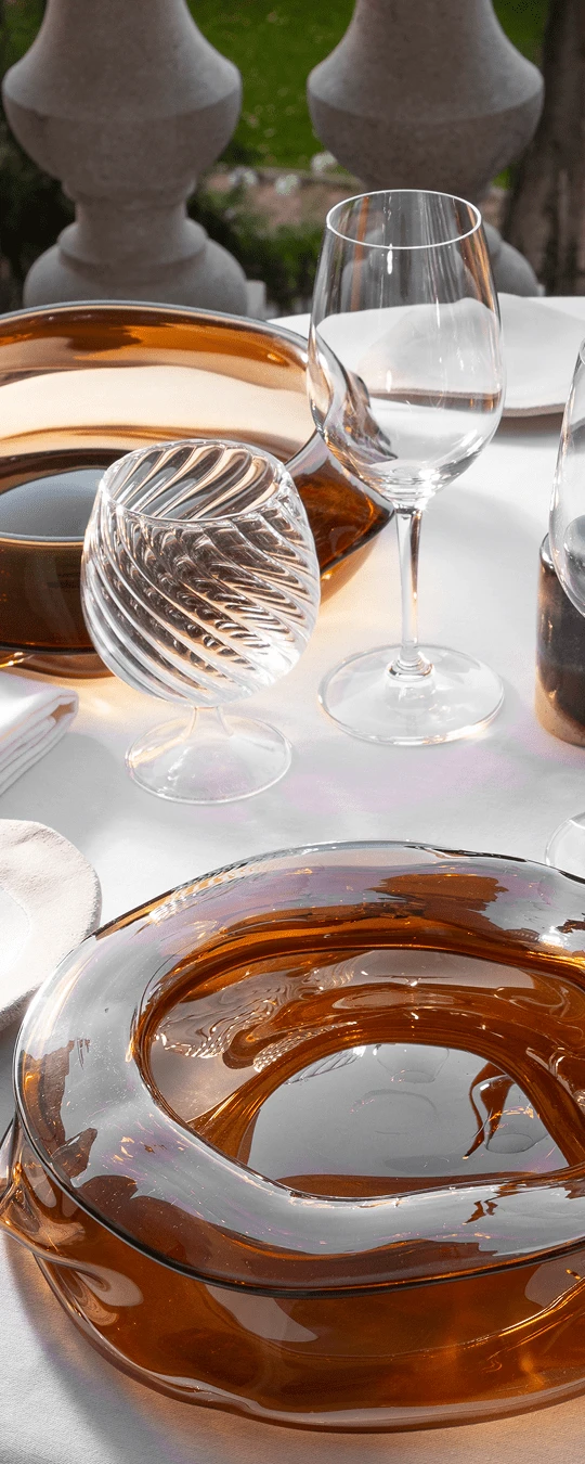 Close-up of a white dining table set with amber glass bowls, a spiral-patterned glass, a wine glass, and plates in an outdoor setting.