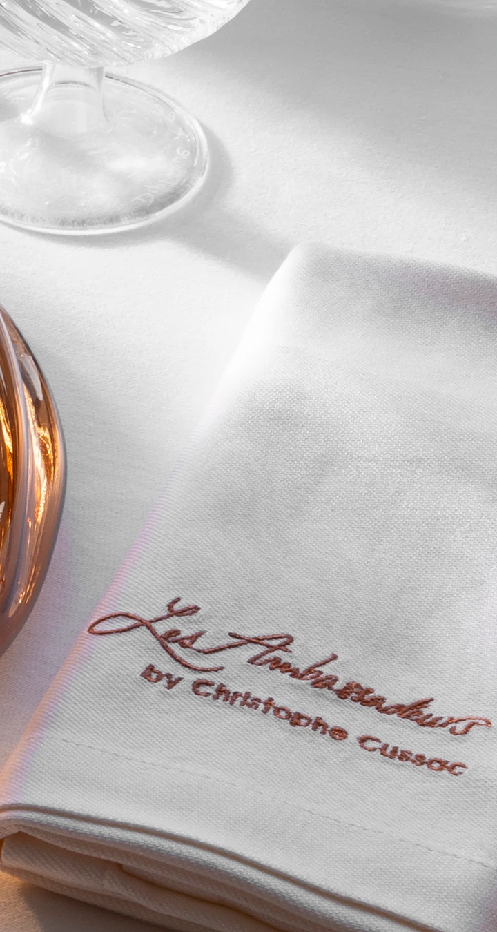 White cloth napkin embroidered with 'Les Ambassadeurs by Christophe Cussac' placed on a white table next to the base of a glass and a reflective surface.