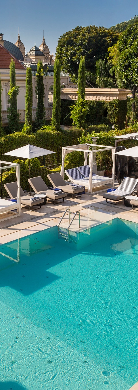 Outdoor swimming pool with clear blue water surrounded by sun loungers, white umbrellas, and cabanas next to a hedge and tall trees.