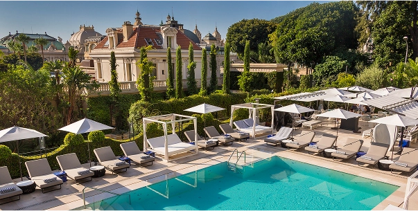 Outdoor swimming pool surrounded by lounge chairs, cabanas, and umbrellas with a historic building and green trees in the background under a clear sky.