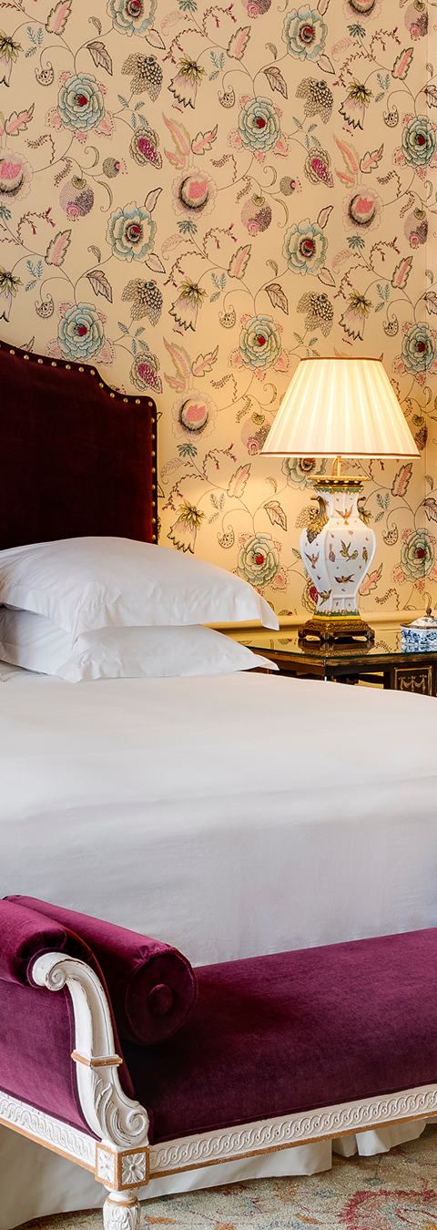 Elegant bedroom corner with white bed linens, a purple velvet bench, floral wallpaper, and a decorative table lamp.