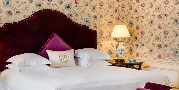 Luxurious hotel room bed with a burgundy velvet headboard, white linens, decorative pillows, floral wallpaper, and a bedside table with an ornate lamp.