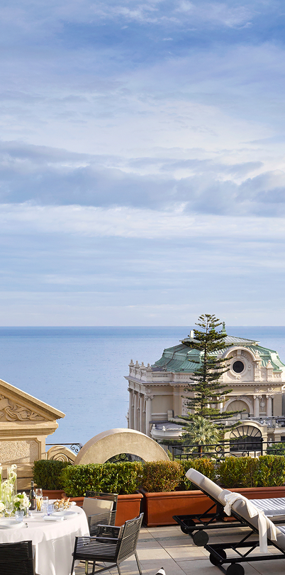 Elegant outdoor terrace with white dining table, chairs, sun loungers, and a backdrop of historic architecture and calm sea under a partly cloudy sky.
