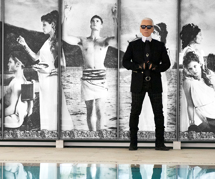 Man in black suit and sunglasses standing in front of large black-and-white classical-style artwork featuring multiple figures by a body of water.