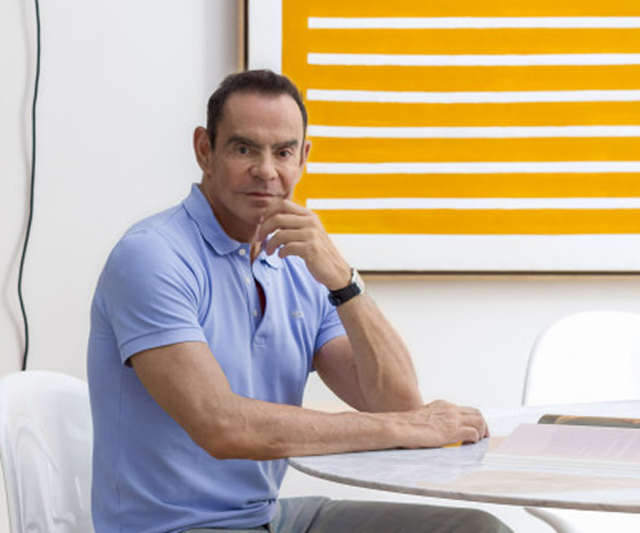 Man in a light blue polo shirt sitting at a white marble table with his hand resting on his chin, with a yellow and white striped artwork on the wall behind him.