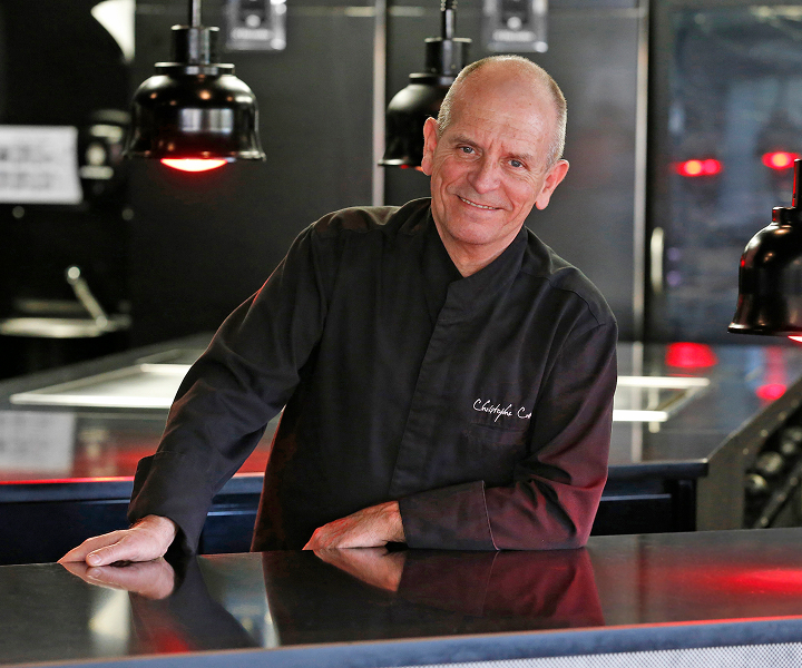 Smiling chef in black uniform leaning on a counter in a modern kitchen with hanging lights.