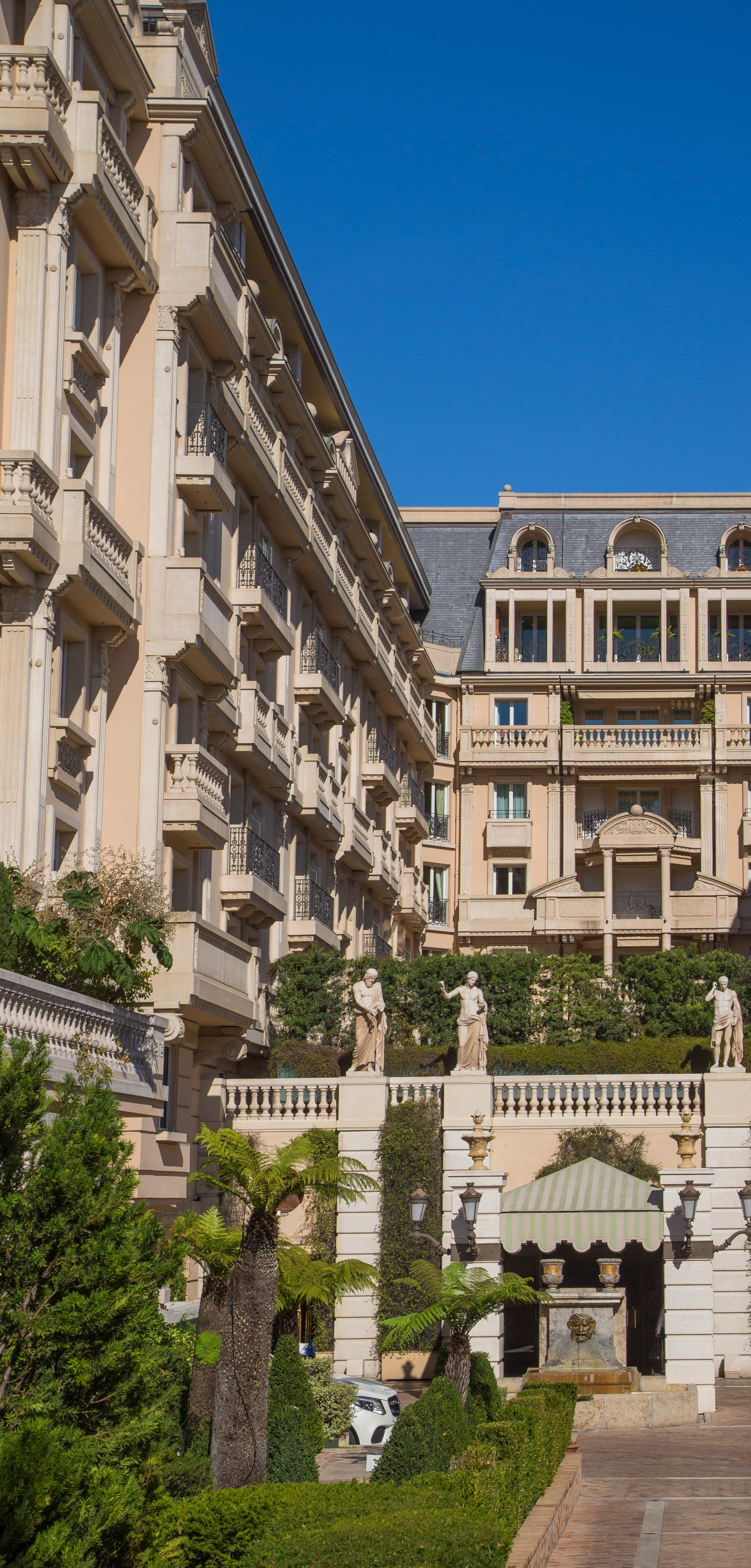 Ornate beige building with multiple balconies, classical statues on a terrace, and a garden with palm trees and shrubs under a clear blue sky.