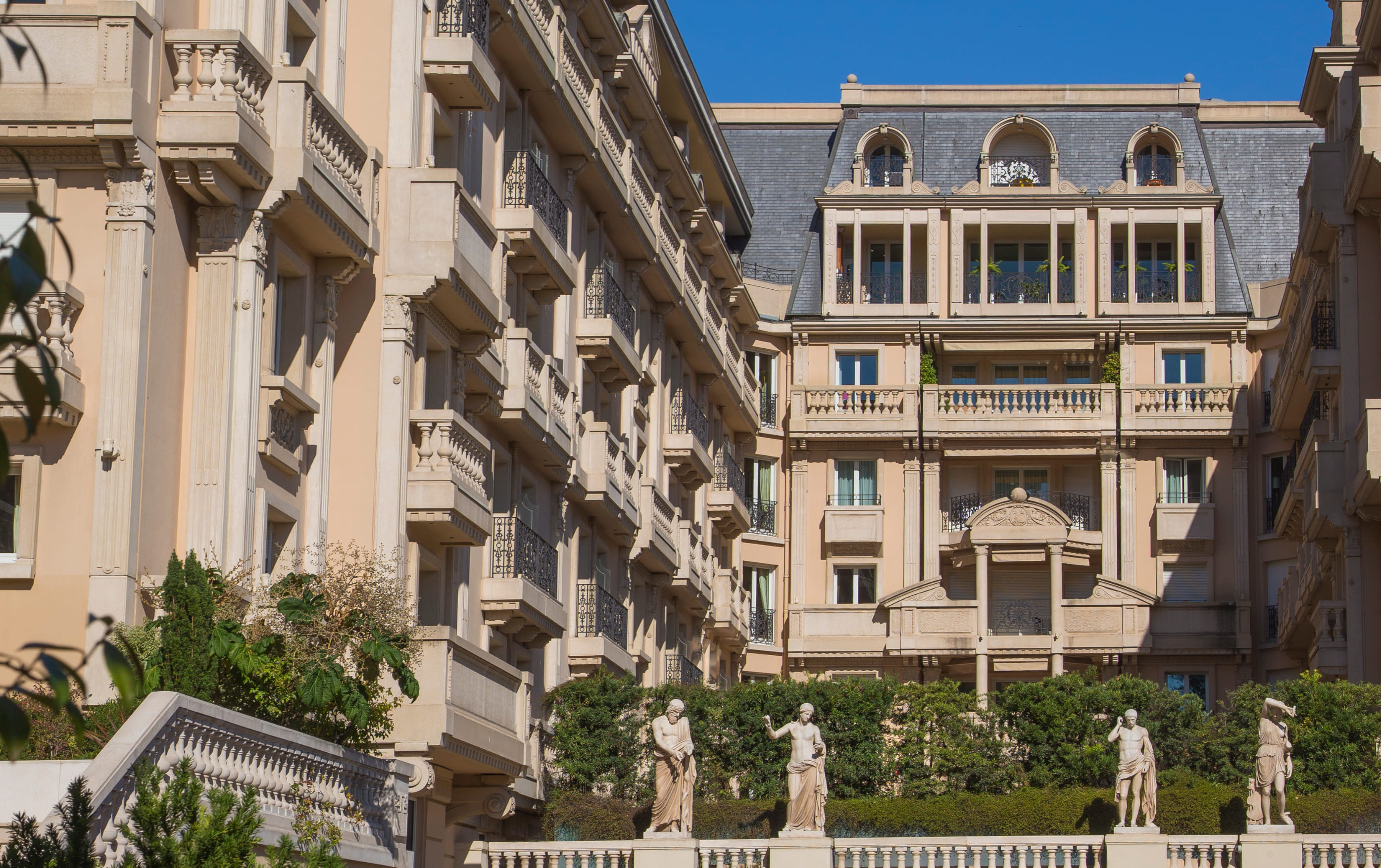 Elegant multi-story beige building with ornate balconies and classical statues in front under clear blue sky.