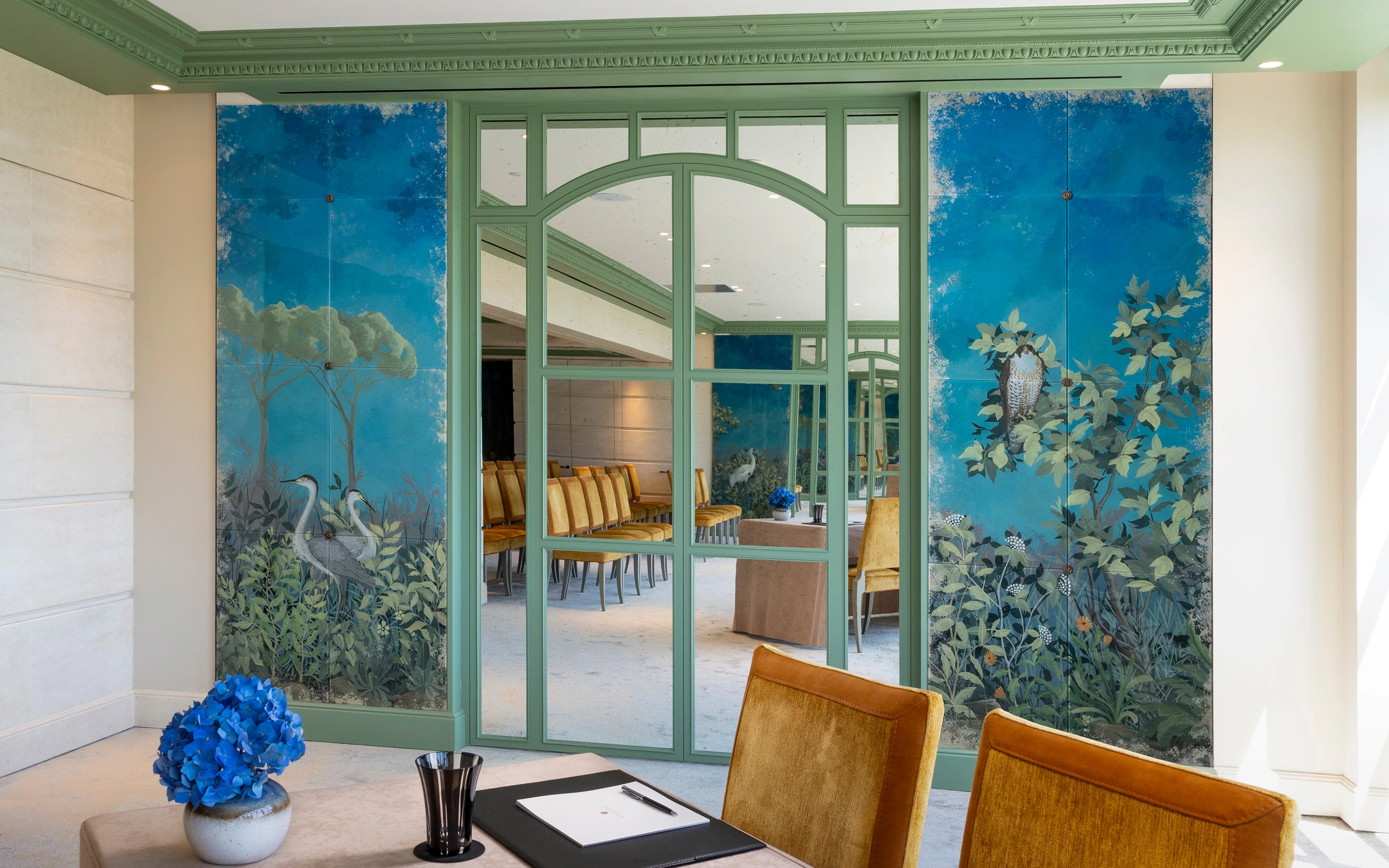 Elegant room with mustard yellow chairs, a table with a blue flower vase, and mirrored green-framed windows flanked by blue nature-themed murals depicting birds and foliage.