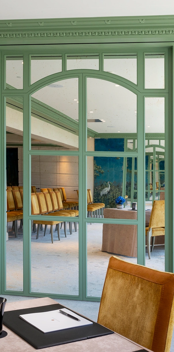 Conference room with mustard velvet chairs reflected in green-framed decorative glass panels, a table with a blue flower centerpiece, and a notepad with pen on a desk in the foreground.