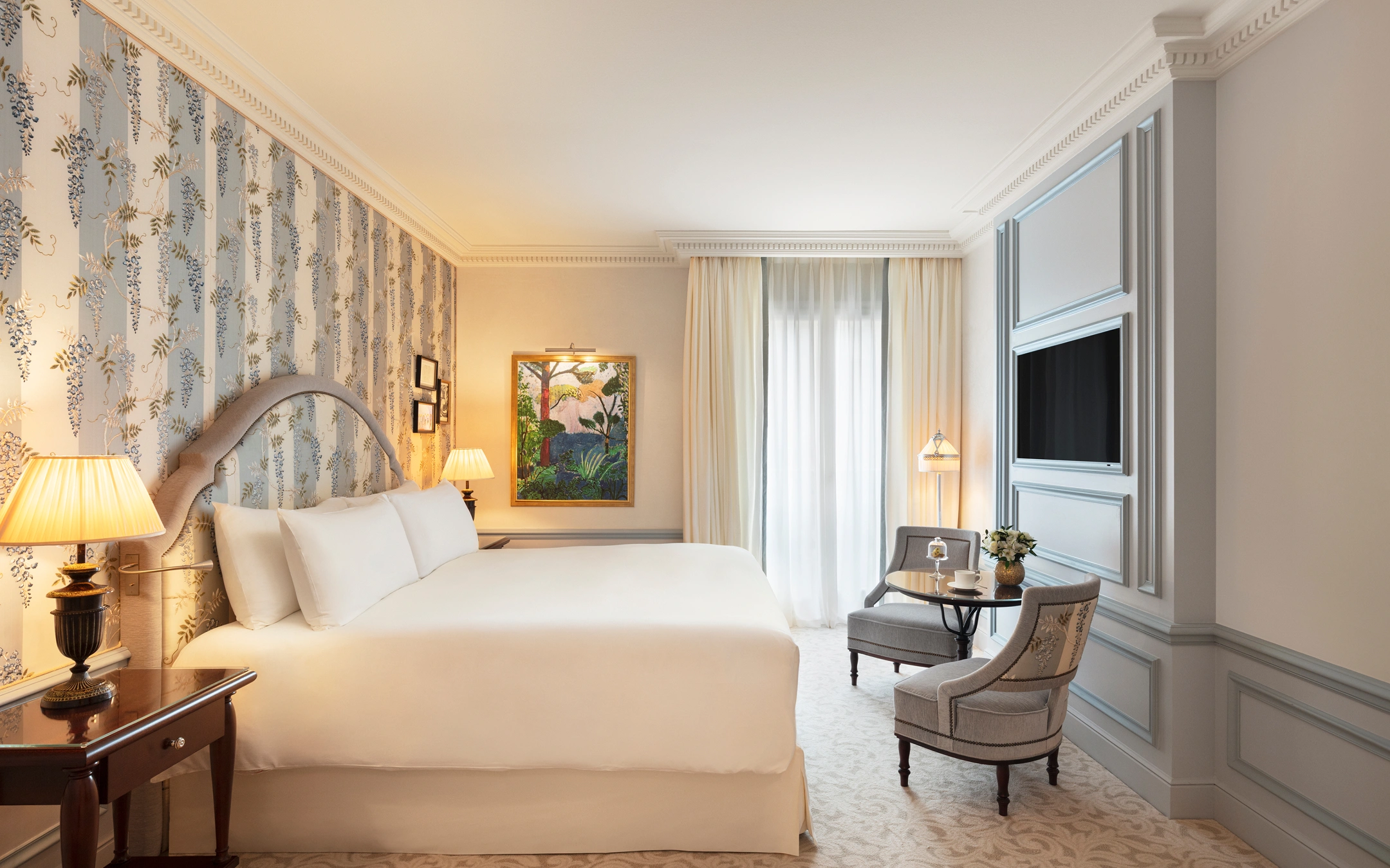 Elegant hotel room with a large white bed, floral wallpaper, bedside lamps, a painting, two gray chairs, a small round table, and a wall-mounted TV.