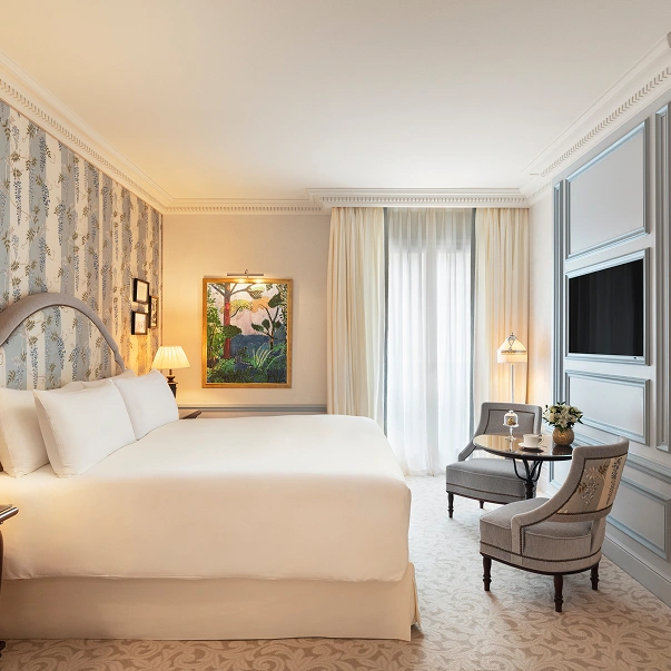 Bright hotel room with a large white bed, floral wallpaper, framed artwork, seating area with two gray chairs, round table, and a mounted flat-screen TV.