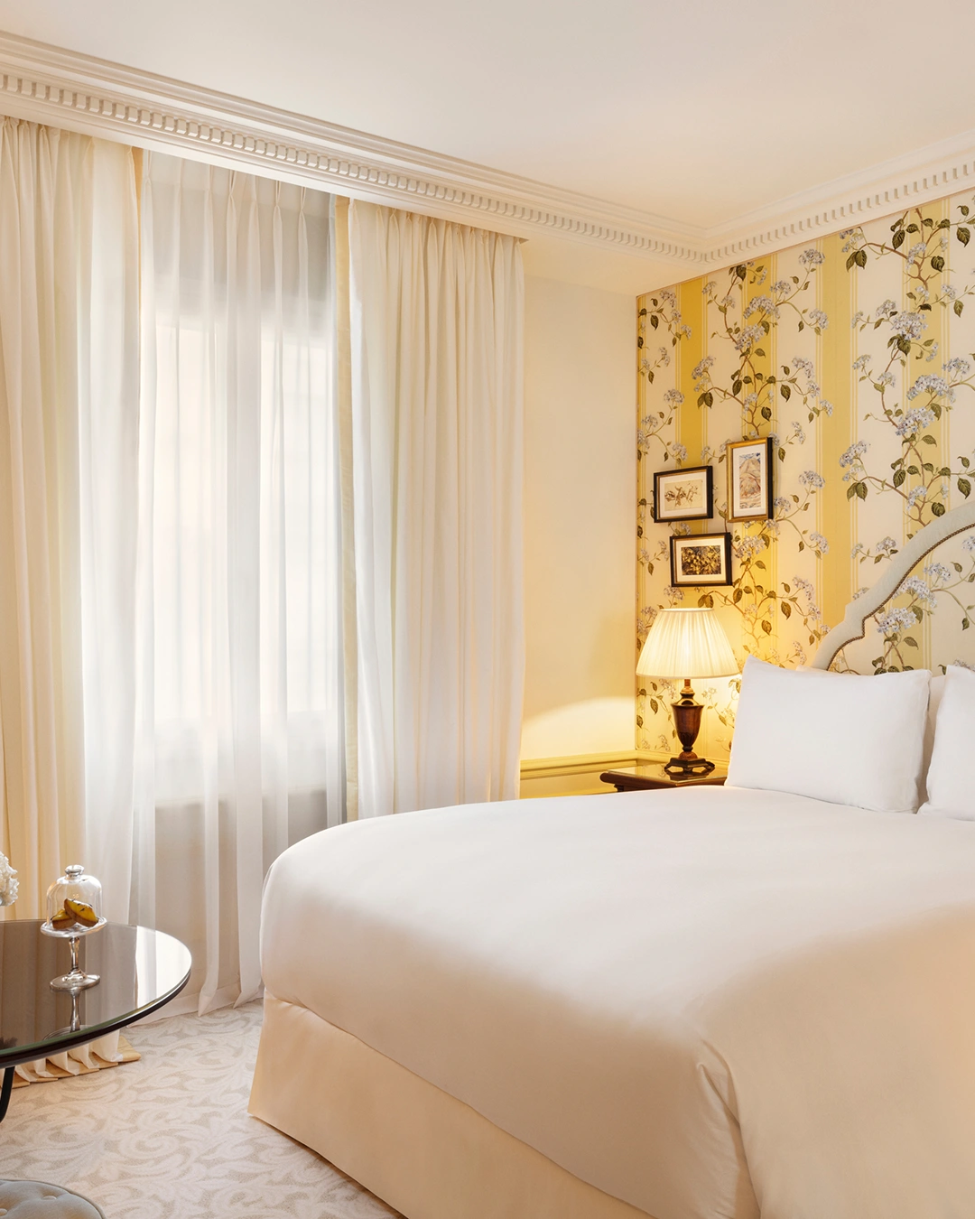 Bright hotel room with a large white bed, floral wallpaper, a bedside table with lamp, and sheer curtains over the window.