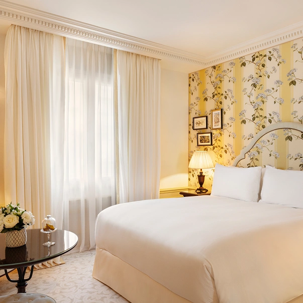 Bright hotel room with a large bed, floral yellow wallpaper, bedside lamp, sheer curtains, and a table with a flower arrangement.