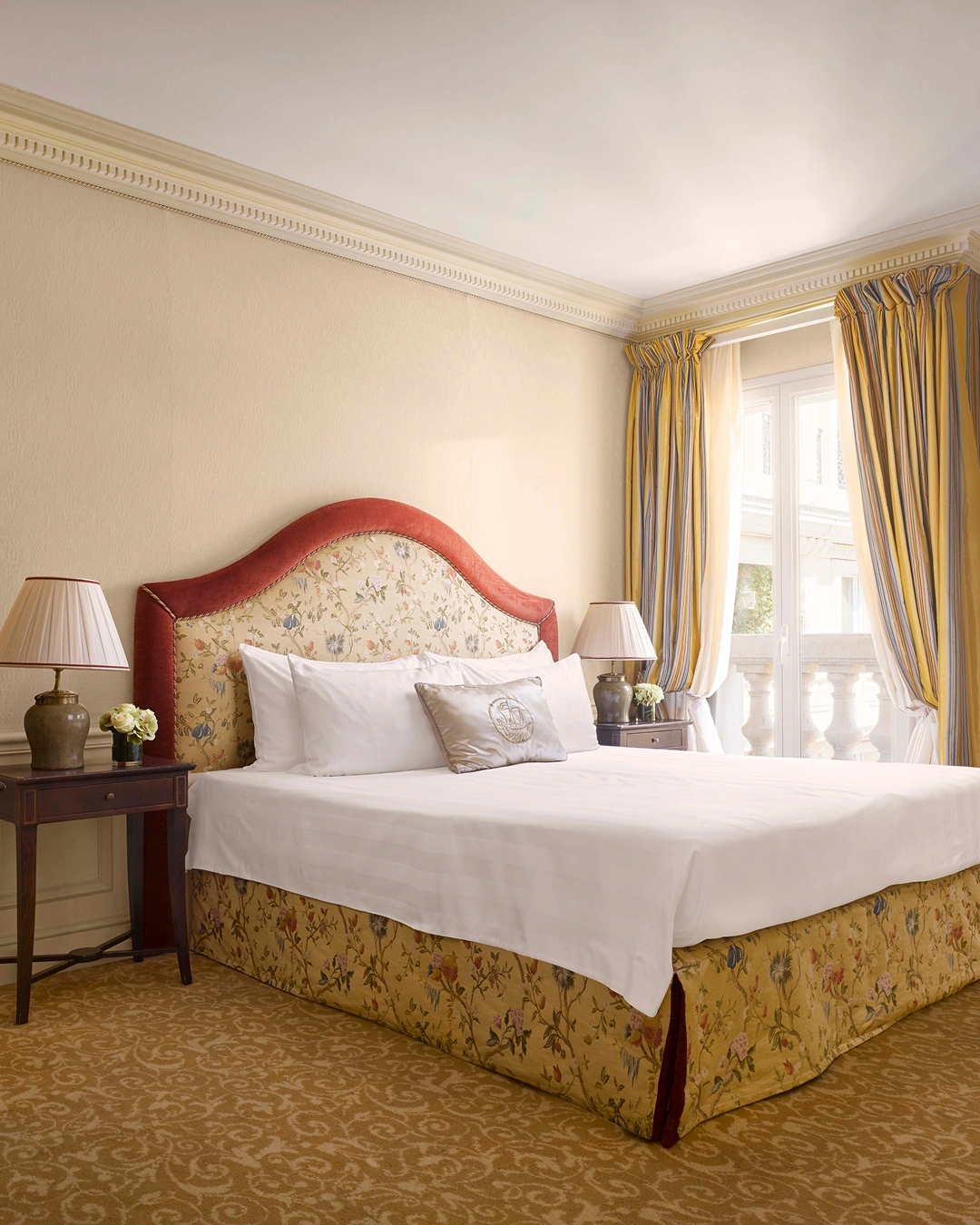 Elegant hotel room with floral patterned bed frame, white linens, bedside tables with lamps, and striped yellow curtains.