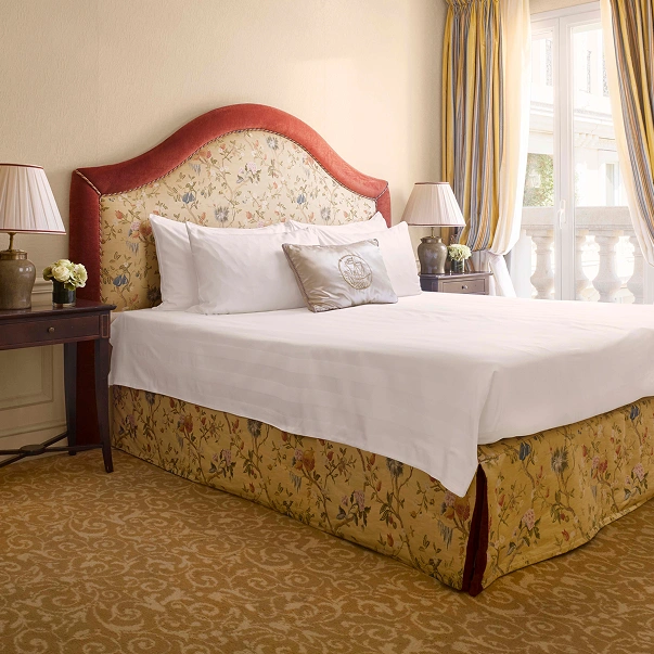Elegant hotel room with a floral patterned headboard and bed skirt, white bedding, decorative pillow, two bedside tables with lamps, and a window with yellow and blue curtains.