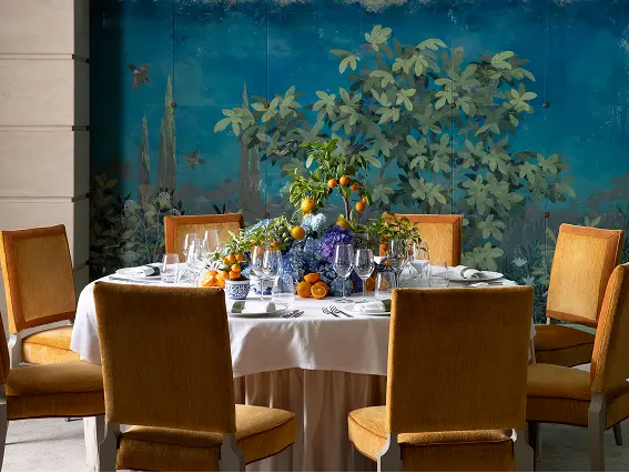 Elegant round dining table set with white tablecloth, glassware, and centerpiece with oranges and flowers, surrounded by six orange upholstered chairs against a blue leafy mural.