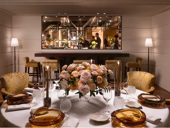 Elegant dining table set with amber glass plates, crystal glasses, candles, and a floral centerpiece in a warmly lit room with a large mirror reflecting people and kitchen lights.