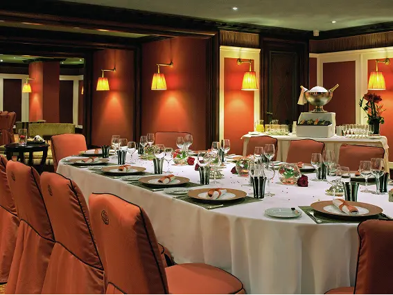 Elegant dining table set with white tablecloth, plates, glasses, and peach-colored chairs in a warmly lit restaurant interior.