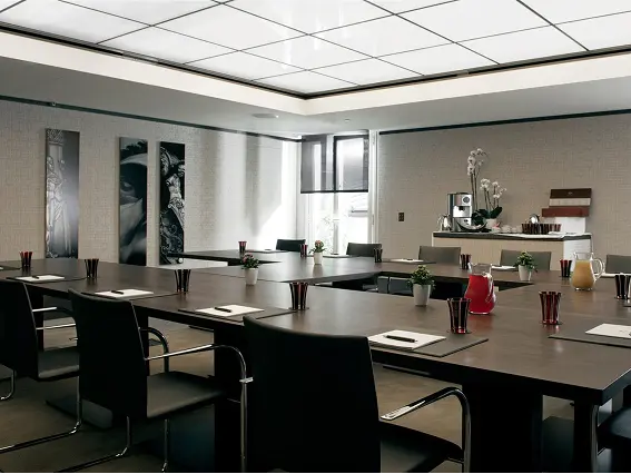 Modern conference room with U-shaped tables, black chairs, notebooks, pencils, plants, and pitchers of beverages.