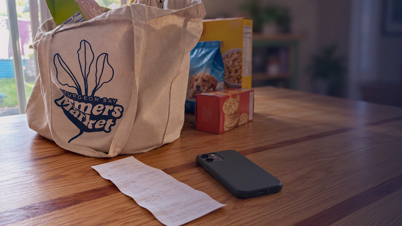 A grocery tote, a box of crackers, dog food, and a cereal box on a dining table, with a paper receipt and a phone beside them