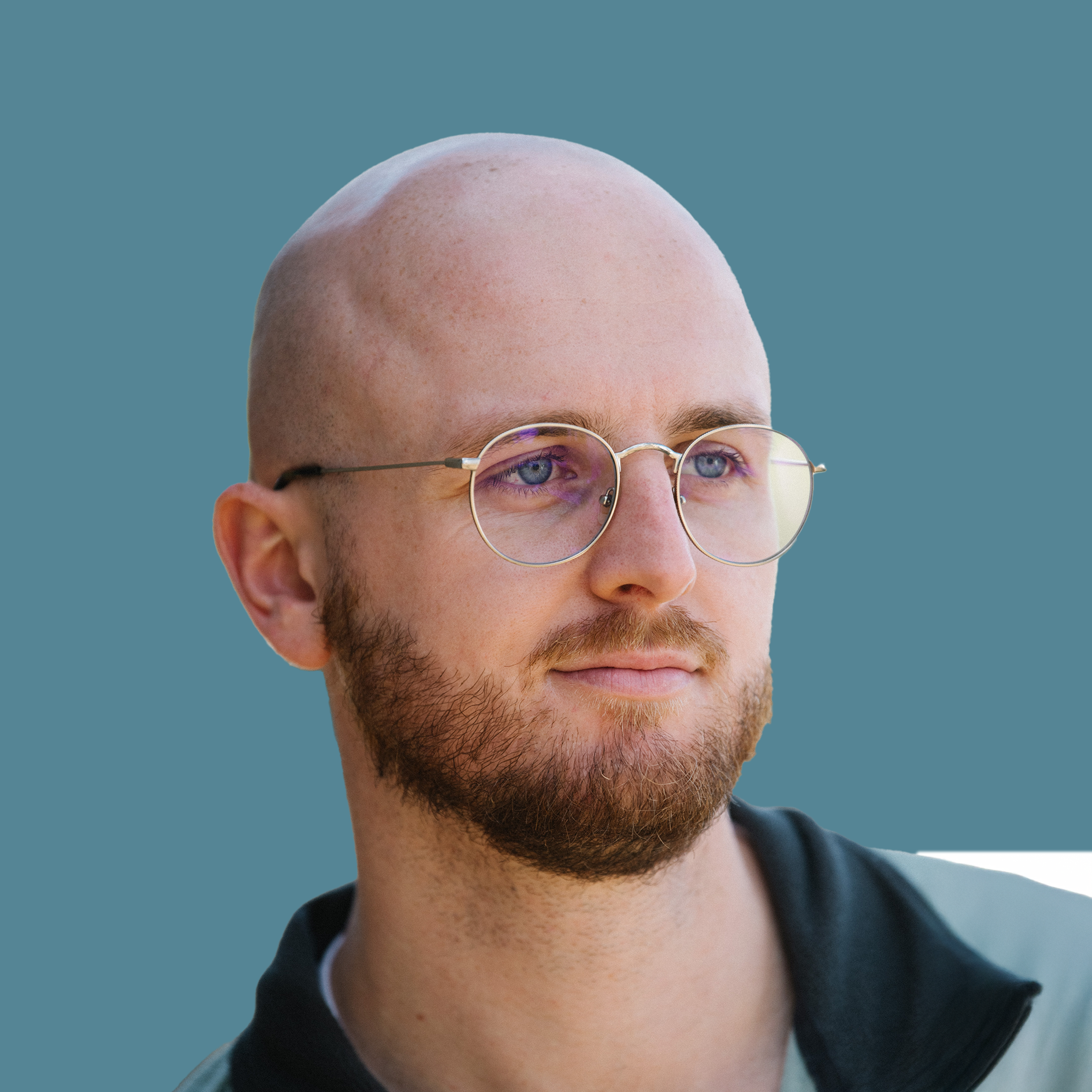 Bald man with a beard and round glasses looking to the right against a plain background.