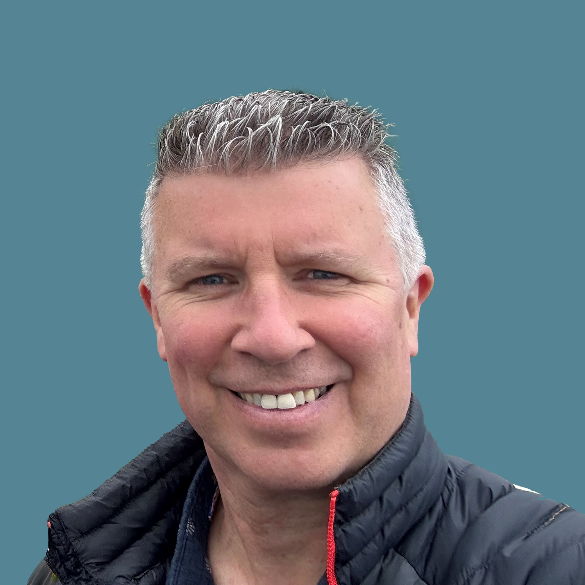 Smiling middle-aged man with short gray hair wearing a black jacket against a plain blue background.