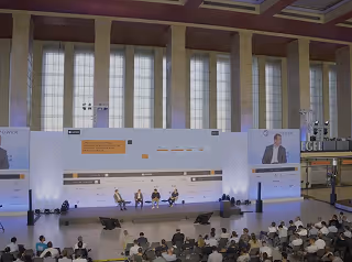 Panel discussion with four speakers seated on stage in a large room with tall windows and an audience watching.