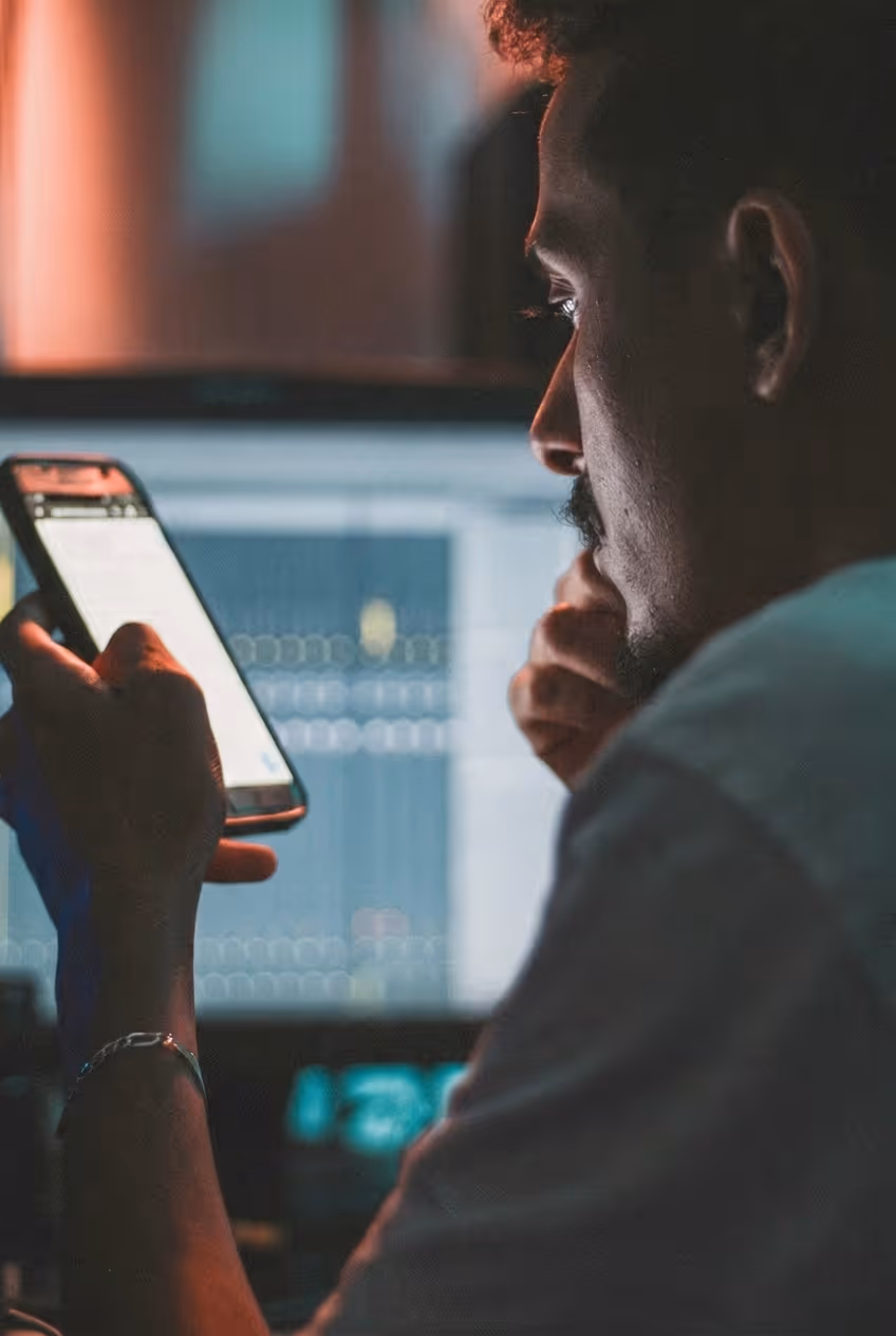 Man intently looking at a smartphone screen in a dimly lit room with a computer monitor in the background.
