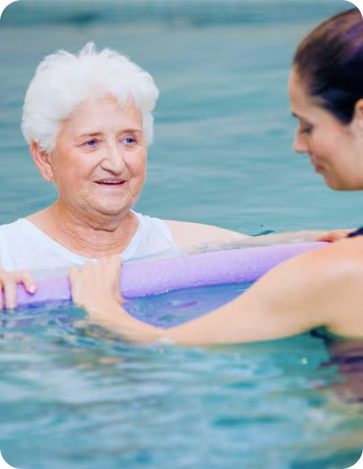Elderly woman in a pool holding a purple floatation bar, assisted by a younger woman in water therapy.