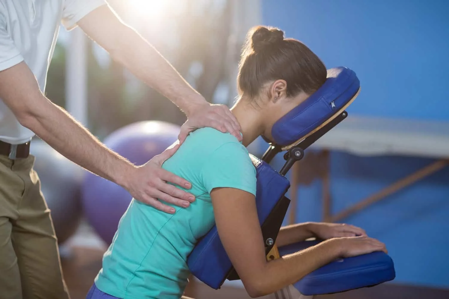 Woman receiving a seated massage on a face cradle chair from a therapist in a bright room.