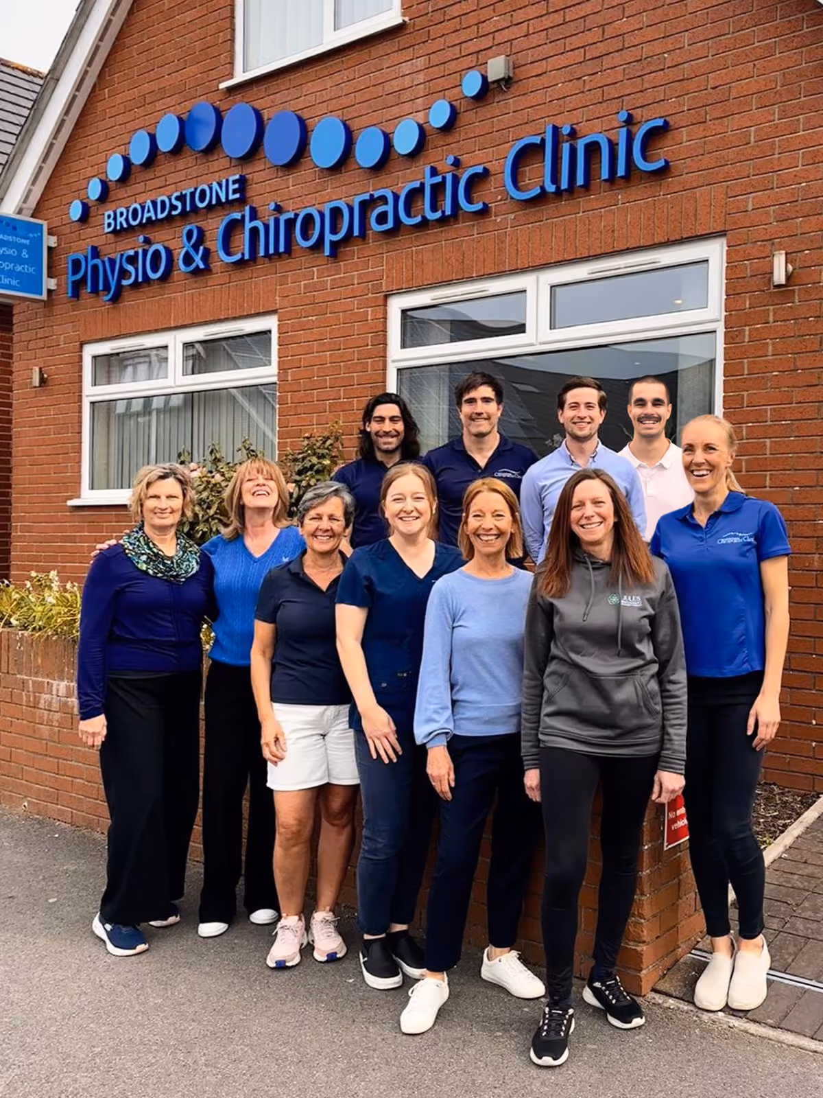Nine staff members stand outside Broadstone Movement Clinic in front of a red brick building with the clinic sign above them.