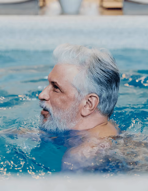 Side view of an older man with gray hair and beard enjoying water in an indoor pool.