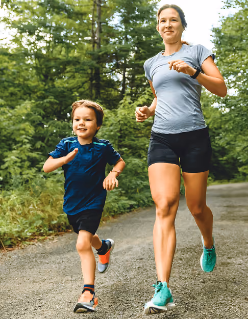 Woman and young boy jogging outside on a forest trail wearing athletic clothes and sneakers.