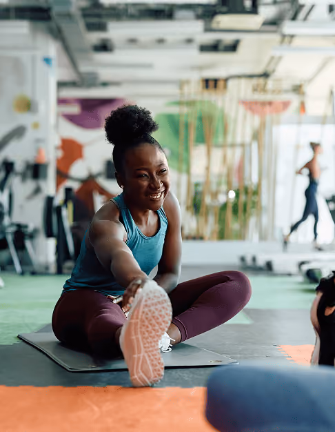 Smiling woman in gym clothes stretching her leg while sitting on an exercise mat indoors.