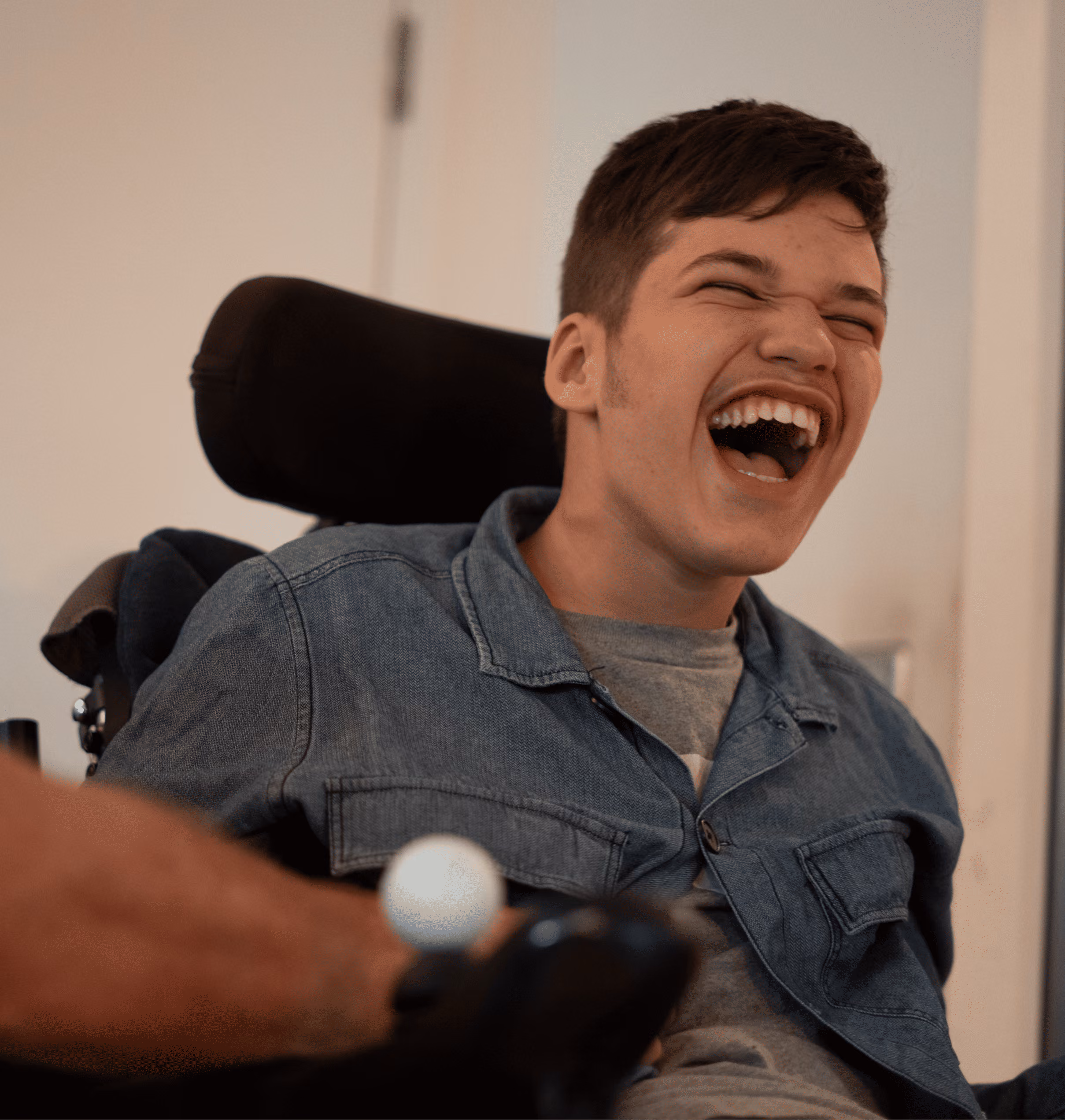 Young man in wheelchair laughing joyfully, wearing a denim jacket and gray shirt indoors.