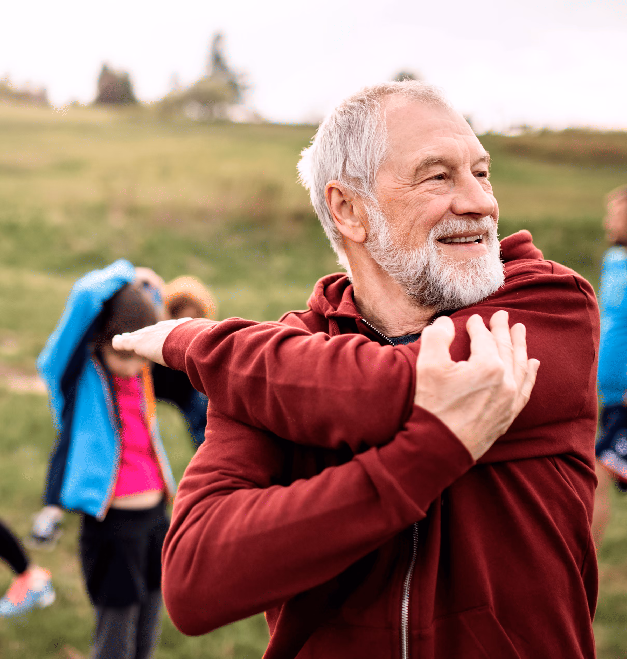 Elderly man in red jacket stretching his arm outdoors with blurred people and greenery in background.