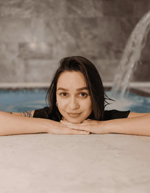 Woman resting her chin on folded hands at the edge of an indoor pool with water flowing in the background.